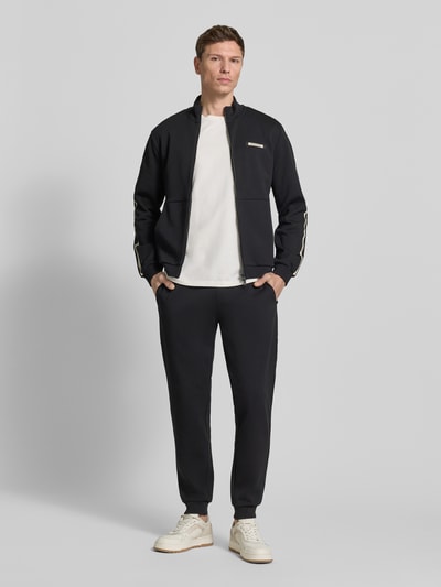 Męski model nosi Bluza rozpinana z stójką i zamek błyskawiczny dwustronny od EA7 Emporio Armani, krój regular fit, wpuszczane kieszenie, detal z logo, długość tyłu 68 cm, długość rękawów 67 cm, ciemny kolor, proste linie, gładka tekstura, całkowity widok z przodu.