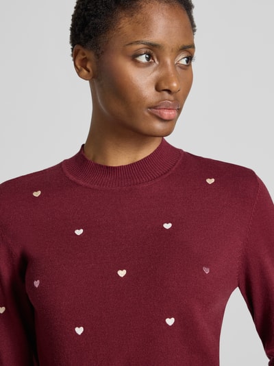 Frauenmodel trägt Strickpullover aus Viskose-Mix mit Stitchings von Apricot in Dunkelrot, kleine Herz-Stickereien, Nahaufnahme Oberkörper. Slim Fit, Rundhalsausschnitt, Allover-Look.