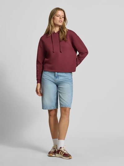 Frauenmodel trägt Regular Fit Hoodie aus Baumwoll-Mix Modell 'CHILLI' von PIECES in Dunkelrot, kombiniert mit hellblauer Jeansshorts, Ganzkörperansicht von vorne. Damen Hoodie.