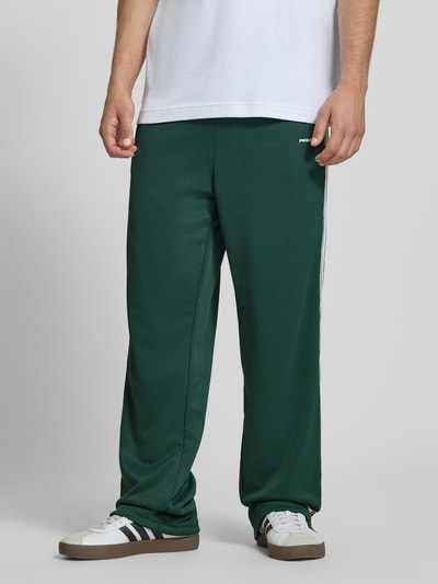 Männermodel trägt Wide Leg Sweatpants mit elastischem Bund von Pegador in Dunkelgrün mit weißem Logo, Material-Mix, Nahaufnahme Oberkörper.