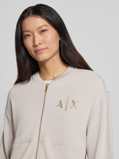 Frauenmodel trägt Sweatjacke mit Label-Print von Armani Exchange in Beige mit goldfarbenem Zweiwege-Reißverschluss, Nahaufnahme Oberkörper. Viskose-Mix mit Stretch-Anteil, lockere Passform, Label-Print, Armani.