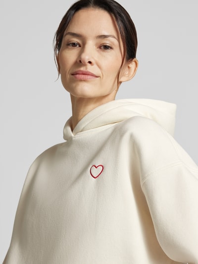 Frauenmodel trägt Hoodie mit Stitching Modell 'Galentine' von OPUS in Creme, Relaxed Fit, sichtbares rotes Herz-Stitching, Nahaufnahme Oberkörper.