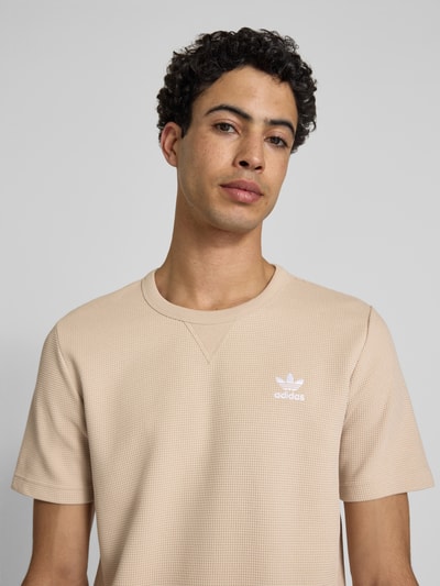 Męski model nosi Regular Fit T-Shirt mit Baumwoll-Anteil von adidas Originals, efekt: jednolity kolor, krój regular fit, wzór: faktura wafla, okrągły dekolt, wyhaftowane logo, rękaw: krótki rękaw, kolor beżowy, fakturowana powierzchnia. Nahaufnahme górnej części ciała.