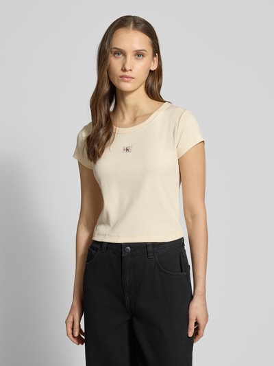 Modelka kobieca ma na sobie Cropped T-Shirt in Ripp-Optik mit Label-Badge od Calvin Klein Jeans, bawełna z elastanem, kremowy, faktura prążkowana, znaczek z logo, długość tyłu w rozmiarze S: 36 cm, widok całej sylwetki z przodu.