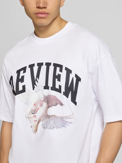 Männermodel trägt T-Shirt mit Logo- und Motiv-Print von REVIEW in Weiß aus Reine Baumwolle, Loose-Fit, gerippter Rundhalsausschnitt, Nahaufnahme Oberkörper. Rückenlänge bei Größe M: 70 cm.