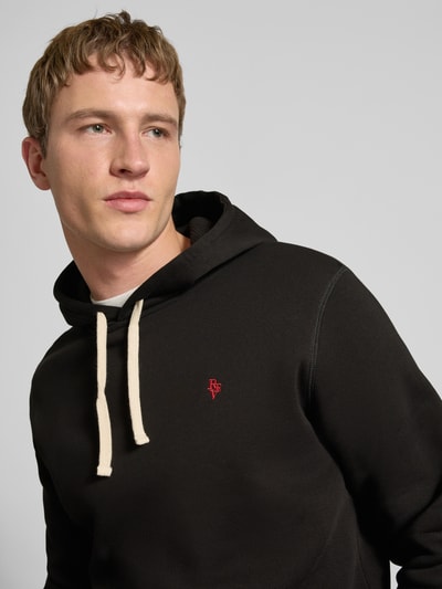 Männermodel trägt Hoodie mit Känguru-Tasche von REVIEW in Schwarz mit Kapuze, Tunnelzug und elastischen Abschlüssen aus Baumwollmischung, Nahaufnahme Oberkörper. Slim-Fit, Rückenlänge bei Größe M 66 cm, Ärmellänge 64 cm.