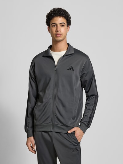 Männermodel trägt Slim Fit Trainingsanzug mit Logo-Stitching von adidas Sportswear in Dunkelgrau mit Stehkragen, Reißverschluss, Eingrifftaschen, elastische Abschlüsse, Ganzkörperansicht von vorne.
