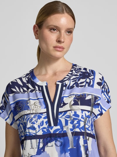 Frauenmodel trägt Blusenshirt aus Viskose mit Strasssteinbesatz von Betty Barclay mit Straight Fit, Tunikakragen, Kappärmel, Motiv- und Statement-Print in Blau- und Weißtönen, Ganzkörperansicht von vorne. Rückenlänge 55 cm, Ärmellänge 19 cm bei Größe 36.