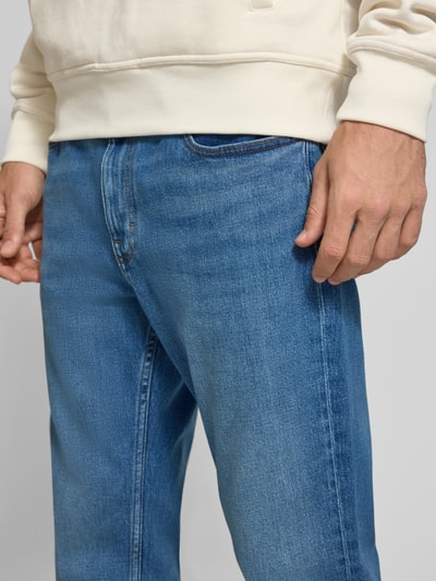 Männermodel trägt Slim Straight Fit Jeans aus Baumwoll-Mix Modell 'RYAN' von Tommy Jeans in Blau mit Kontrastnähten, Nahaufnahme Oberkörper. Denim-Optik, 5-Pocket-Design, Label Patch, Logo-Stitching, Gürtelschlaufen, mittlere Bundhöhe, Knopf mit verdecktem Reißverschluss, Twill-Struktur.