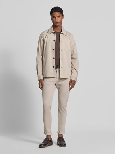 Männermodel trägt Slim Fit Hemdjacke mit Viskose-Anteil von Lindbergh in Beige, elastischer Stoff, Brusttasche, Ganzkörperansicht von vorne. Rückenlänge Größe M: 70 cm, Umlegekragen, durchgehende Knopfleiste, Eingrifftaschen.