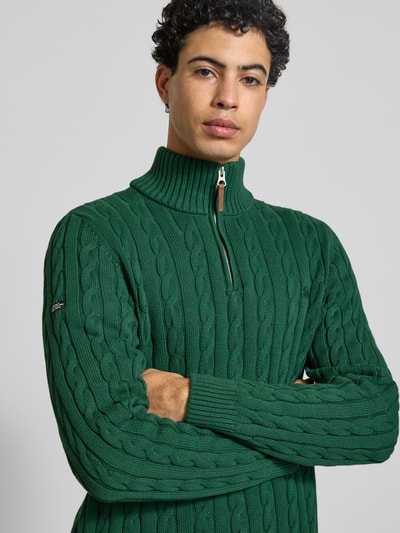 Mannelijk model draagt gebreide pullover uit puur katoen met schipperskraag, kabelpatroon, labeldetail, ribboorden, groene kleur, Strickpullover aus reiner Baumwolle van Superdry, eng aanliggende pasvorm, ruglengte maat M 70 cm, mouwlengte 71 cm, nahaufnahme bovenlichaam.