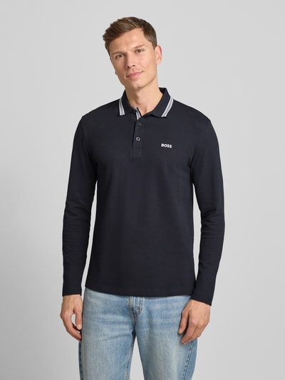 Męski model nosi Regular Fit Koszulkę polo z czystej bawełny Model 'PLISY' od BOSS Green, efekt: jednolity kolor, aplikacja z wyhaftowanym logo, kołnierzyk polo, dekolt henley, listwa guzikowa, rękawy: długi rękaw, wykończenie materiału: pika, kolor ciemny, przód całej sylwetki.