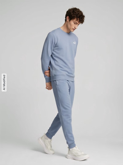 Männermodel trägt hellblaue Jogginghose und Sweatshirt, dazu weiße Sneaker, neutraler Hintergrund, Ganzkörperansicht von vorne, KI-modifiziertes Bild.