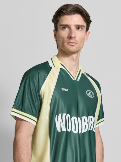 Männermodel trägt Poloshirt mit Label-Print Modell 'Taka' von WOODBIRD, grün-gelb, lockere Passform, kurzärmelig, Nahaufnahme Oberkörper