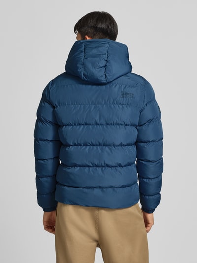 Männermodel trägt Steppjacke mit Label-Stitching und Kapuze von Superdry in Dunkelblau, gesteppte Struktur, Ganzkörperansicht Rückansicht. Herren Steppjacke, gerader Schnitt, Kapuze mit Kordelzug, Reißverschluss, Label-Stitching, seitliche Eingrifftaschen, elastische Ärmelabschlüsse, Rückenlänge 67 cm, Schulterbreite 48 cm, Brustweite 58 cm, Ärmellänge 72 cm.