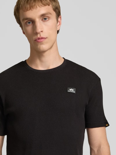 Männermodel trägt T-Shirt mit Label-Patch von Alpha Industries in Schwarz, Reine Baumwolle, Körpernahe Passform, Rundhalsausschnitt, Nahaufnahme Oberkörper.