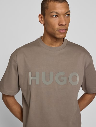 Männermodel trägt Relaxed Fit T-Shirt aus reiner Baumwolle Modell 'DINKEE' von HUGO in Taupe mit 3D Label Print, Nahaufnahme Oberkörper.