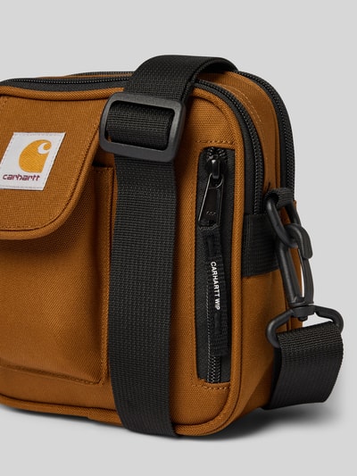Produktfoto, Crossbody Bag mit Logo-Patch von Carhartt Work In Progress in bruin met zwarte verstelbare schouderband, hoofdvak met ritssluiting, voorvak, logopatch, afneembare schouderband, breedte 17 cm, lengte 18 cm, diepte 6 cm, bandlengte 80 cm, textuur van canvas
