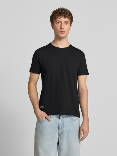 Männermodel trägt T-Shirt mit Label-Patch von Alpha Industries in Schwarz, Reine Baumwolle, Gerader-Schnitt, Rundhalsausschnitt, Ganzkörperansicht von vorne. Rückenlänge bei Größe M: 66 cm.