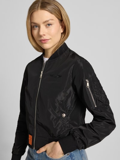 Vrouwenmodel draagt Bomberjacke mit Label-Patch und Reißverschlusstasche am Ärmel van Original Bombers, zwarte kleur, glanzend oppervlak, rits, mouwzak, logo-stiksel, opstaande kraag, zijzakken met drukknoop, elastische boorden, relaxed fit, voorkant volledig zichtbaar, heuplengte, nahaufnahme bovenlichaam.
