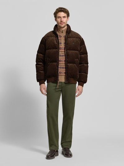 Männermodel trägt Regular Fit Bomberjacke aus reiner Baumwolle von Polo Ralph Lauren in Dunkelbraun, kombiniert mit gemustertem Pullover und grüner Hose, Ganzkörperansicht von vorne, Herren Bomberjacke / Blouson von Polo Ralph Lauren.