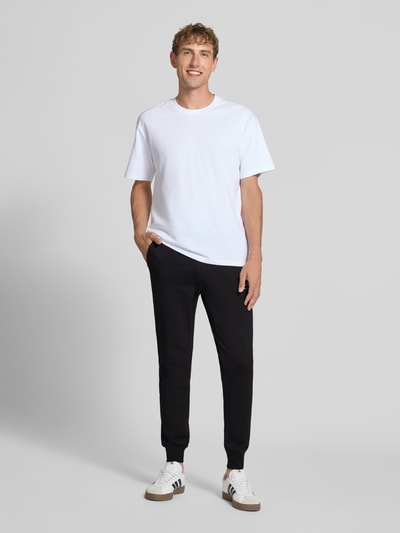 Męski model nosi spodnie dresowe z nadrukiem z logo Model 'GORDON' od JACK & JONES, czarne, luźny krój, wpuszczane kieszenie, pas z tunelem, długość wewnętrzna nogawki w rozmiarze M: 77 cm, widok całej sylwetki z przodu.