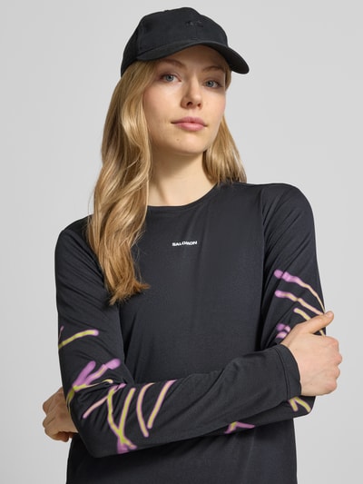 Kobiece modelka nosi Funktionsshirt mit Logo Modell 'Kout' od Salomon, krój regular fit, okrągły dekolt, czarny kolor z kolorowymi wzorami na rękawach, długość pleców w rozmiarze S: 58 cm, zbliżenie na górną część ciała