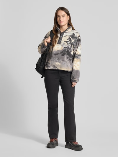 Modelka kobieca ma na sobie Regular Fit Pullover mit Logo-Patch Modell 'Helvetia' od COLUMBIA w odcieniach szarości, kremu i czerni, z teksturą polaru, kieszenią na piersi, elastycznymi mankietami, stójką oraz zapięciem na guziki, kontekst: damski sweter, krój regular fit, poliestrowa struktura, szerokość w rozmiarze S: 58 cm, długość tyłu: 55 cm, szerokość ramion: 44 cm, długość rękawa: 58 cm; widok całej sylwetki z przodu.