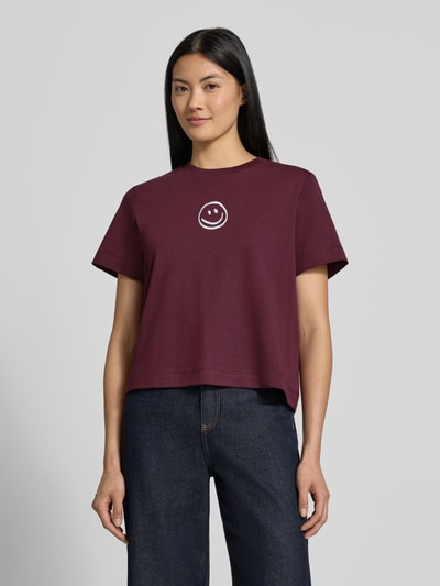 Kobiece model nosi damski t-shirt Boxy Fit T-Shirt aus reiner Baumwolle model 'SARA' od VERO MODA w kolorze bordowym z białym nadrukiem, prosta forma, gładka tekstura, widok całej sylwetki z przodu.