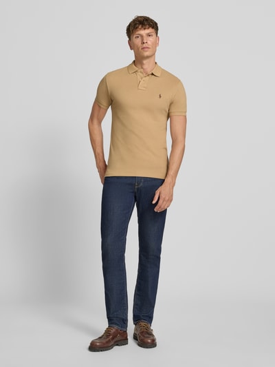 Męski model nosi Slim Fit Poloshirt aus reiner Baumwolle von Polo Ralph Lauren, beżowa koszulka polo, granatowe spodnie, brązowe buty, prosty krój, gładka faktura, Ganzkörperansicht von vorne.