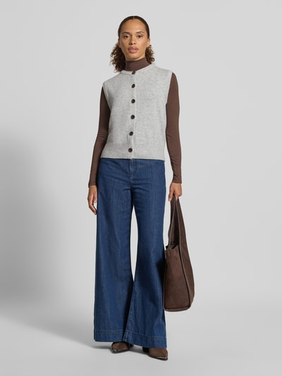 Frauenmodel trägt Regular Fit Strickweste aus Woll-Mix von Christian Berg, grau mit Knopfleiste, gerippte Abschlüsse, ärmellos, kombiniert mit braunem Langarmshirt und blauer weiter Jeans, Produktlänge 45 cm bei Größe S, Ganzkörperansicht von vorne.