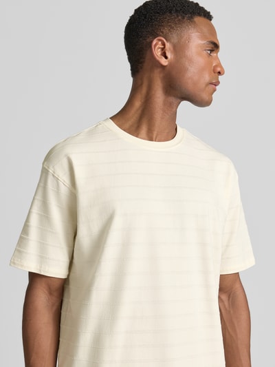 Männermodel trägt T-Shirt mit Strukturmuster und Rundhalsausschnitt von Scotch & Soda, Cremefarbe, Baumwoll-Elasthan-Mix, Relaxed-Fit, Nahaufnahme Oberkörper. Rückenlänge bei Größe M: 72 cm.