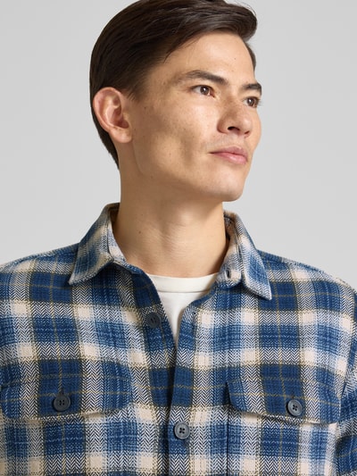 Męskki model nosi Relaxed Fit Freizeithemd mit Brusttaschen od Fynch-Hatton z bawełny, krój relaxed fit, kołnierzyk kent, sportowe mankiety, koszula casualowa w niebieską kratę, z długim rękawem, listwa guzikowa, kieszenie na piersi, długość tyłu 78 cm dla rozmiaru M, zbliżenie na górną część ciała.