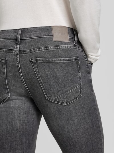 Männermodel trägt Modern Fit Jeans aus Baumwoll-Mix Modell 'Chuck' von Brax, stylizowana na denim, detal z logo, 5 kieszeni, splot twill, szlufki na pasek, zapięcie na guzik i zamek błyskawiczny, widoczna tekstura materiału, szarość i biel, widok z tyłu, detal zbliżeniowy.