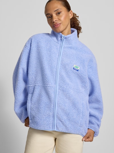 Frauenmodel trägt Regular Fit Jacke aus Teddyfell Modell 'Hoktown' von American Vintage, hellblau, Stehkragen, Logo-Patch, Reißverschluss, Eingrifftaschen, grob strukturierte Oberfläche, Ganzkörperansicht von vorne.