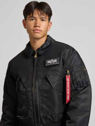 Männermodel trägt Bomberjacke mit seitlichen Pattentaschen und Label-Patch von Alpha Industries, Schwarz mit Reißverschlüssen, Nahaufnahme Oberkörper. Herrenmodell Regular Fit mit Kentkragen, Reißverschlusstasche am Ärmel und Label-Detail, gerippte Abschlüsse.