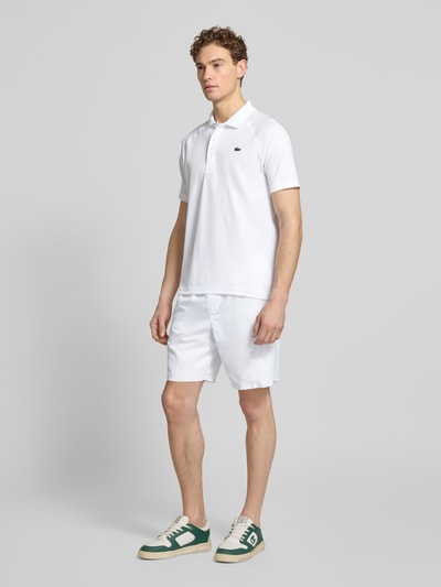 Męski model nosi Regular Fit Sport Poloshirt mit Label Patch od Lacoste Sport, biały kolor, krój regular fit, piqué, krótki rękaw, unikatowe detale, widok całej sylwetki z przodu.