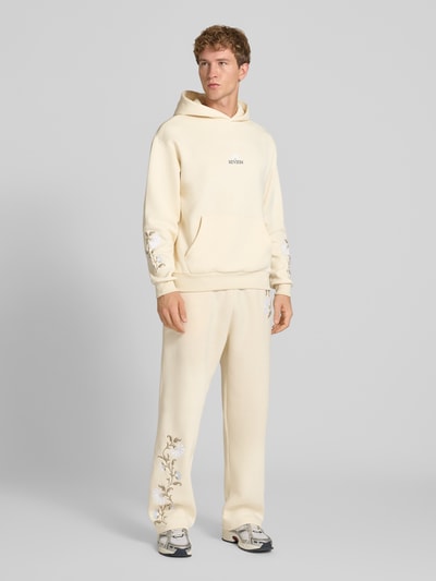 Männermodel trägt Relaxed Fit Hoodie mit Känguru-Tasche von REVIEW in Creme mit floraler Stickerei, elastische Abschlüsse, Ganzkörperansicht von vorne.