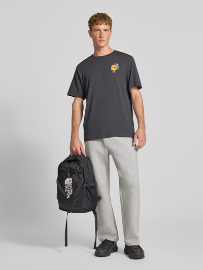 Männermodel trägt T-Shirt aus reiner Baumwolle Modell 'Gear Up Get Out' von The North Face in Dunkelgrau, dezenter Logo-Print, Regular Fit, Kurzarm, Rundhalsausschnitt, kombiniert mit hellgrauer Hose, Ganzkörperansicht von vorne.