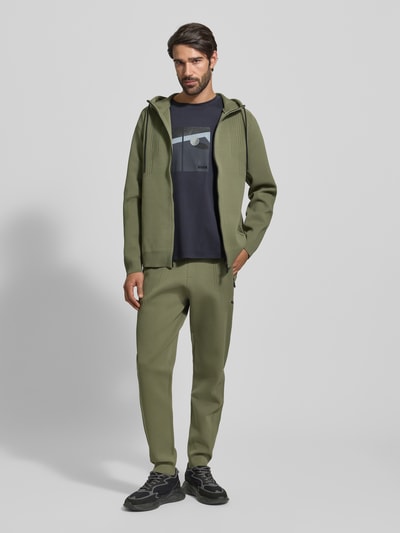 Männermodel draagt Regular Fit Trainingsjacke mit Jacquard Elementen Modell 'ACTIVE PUSH' van BOSS Green in effen olijfgroen met gestructureerde stof, waterdichte ritssluiting, label-applicatie en geribde boorden, vooraanzicht hele lichaam.