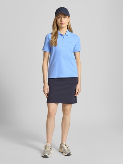 Frauenmodel trägt Poloshirt mit Logo-Stitching und Polokragen von Gant in Hellblau, Reine Baumwolle, Regular-Fit, kurze Knopfleiste, Rückenlänge bei Größe S: 57 cm, kombiniert mit dunklem Rock, Sneakers und Basecap, Ganzkörperansicht von vorne.