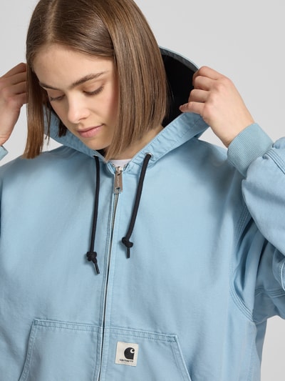 Frauenmodel trägt Sweatjacke mit Kapuze und Känguru-Tasche von Carhartt Work In Progress in Hellblau, Reine Baumwolle, Relaxed-Fit, Label-Detail am Reißverschluss, Nahaufnahme Oberkörper.