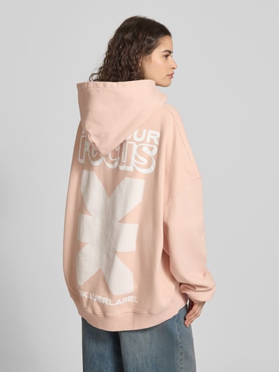 Frauenmodel trägt Oversized Hoodie mit Känguru-Tasche von Karo Kauer in Rosa mit Label-Print aus Reine Baumwolle, Ganzkörperansicht Rückansicht.