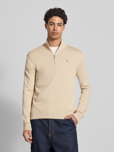 Männermodel trägt Slim Fit Troyer aus reiner Baumwolle von Tommy Jeans in Beige, unifarben, Reißverschluss, Feinstrick, Ganzkörperansicht von vorne.