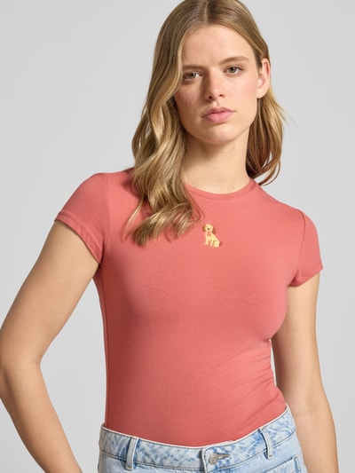 Kobiece model nosi damski t-shirt z mieszanki bawełny i elastanu, krój slim fit, wyhaftowany motyw, okrągły dekolt, długość tyłu 54 cm w rozmiarze S, T-Shirt mit Motiv-Stitching und Rundhalsausschnitt od REVIEW; jasnoróżowy, gładka tekstura, widok na górną część tułowia z przodu.