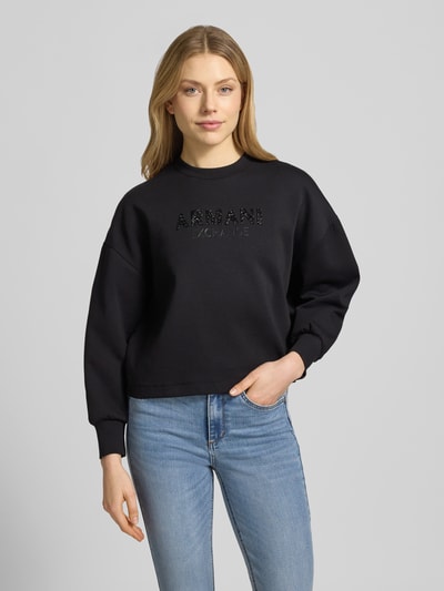 Kobiece model prezentuje Sweatshirt mit Logo-Print und Rundhalsausschnitt von ARMANI EXCHANGE, mieszanka bawełny z dodatkiem streczu, krój loose fit, okrągły dekolt, nadruk z logo, czarny kolor, gładka faktura, widok całej sylwetki z przodu.