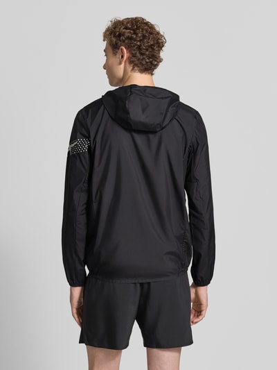 Męski model nosi Regular Fit kurtkę z nadrukiem z logo Model 'ICON' od ASICS TIGER, czarna, kaptur, stójka, długość tyłu 72 cm, długość rękawów 67 cm, elastyczne wykończenia, widok całej sylwetki z tyłu.