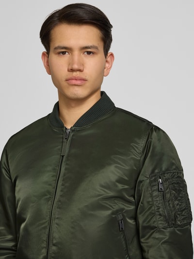 Männermodel trägt Bomberjacke mit Kapuze und Reißverschlusstaschen von Superdry in Grün, Regular-Fit, Nahaufnahme Oberkörper. Rückenlänge 56 cm, Schulterbreite 45 cm, Brustweite 60 cm, Ärmellänge 68 cm, Größe M.