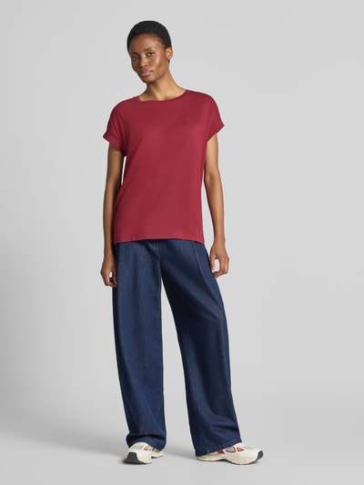 Frauenmodel trägt dunkelrotes Loose Fit T-Shirt aus reiner Baumwolle Modell 'IDAARA' von ARMEDANGELS, kombiniert mit weiter blauer Hose, Ganzkörperansicht von vorne.