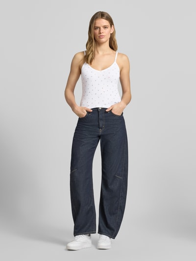 Vrouwenmodel draagt Trägertop met V-hals en logo van Guess Jeans, wit met kleine logo details, materiaalmix met viscose, regular fit, verstelbare spaghettibandjes, ruglengte maat S 28 cm, gecombineerd met donkere jeans; Ganzkörperansicht von vorne.